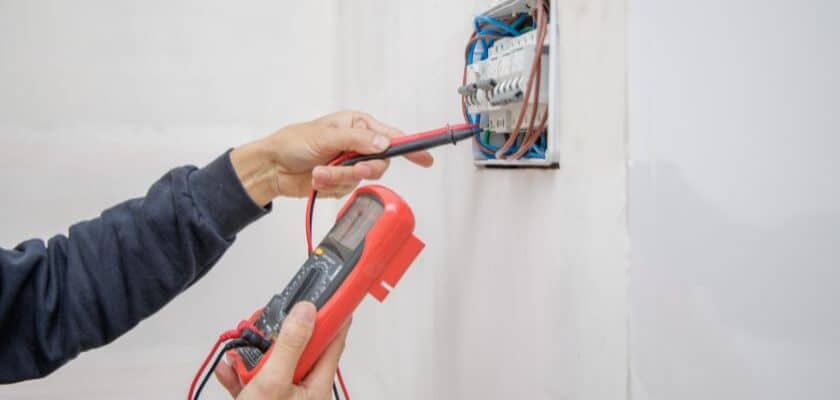 Uma foto de uma mão segurando um voltímetro e a outra segurando uma espécie de chave conectora na parede