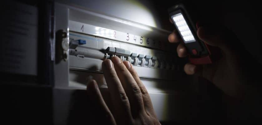 Um ambiente escuro, provavelmente devido a uma falha de energia (blecaute). Uma mão humana está na frente de um quadro de distribuição elétrico (painel de disjuntores), tentando manusear um dos disjuntores. A única fonte de luz visível é o feixe brilhante de uma lanterna LED