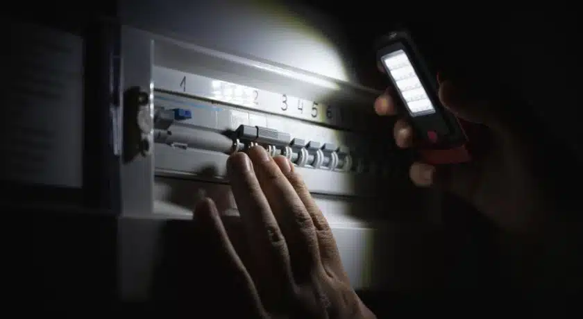 Um ambiente escuro, provavelmente devido a uma falha de energia (blecaute). Uma mão humana está na frente de um quadro de distribuição elétrico (painel de disjuntores), tentando manusear um dos disjuntores. A única fonte de luz visível é o feixe brilhante de uma lanterna LED