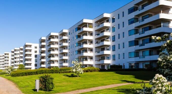 Uma linha de edifícios residenciais modernos, brancos e de múltiplos andares, com várias varandas, estendendo-se sob um céu azul claro. Há um gramado verde bem cuidado e alguns arbustos e árvores floridas