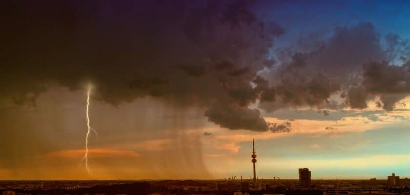 Cenário de uma tempestade severa sobre uma cidade, com nuvens escuras, chuva intensa e um raio vertical atingindo o solo à esquerda. A direita o céu mais limpo com algumas novens