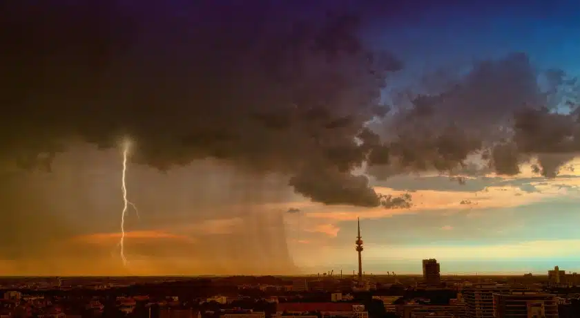 Cenário de uma tempestade severa sobre uma cidade, com nuvens escuras, chuva intensa e um raio vertical atingindo o solo à esquerda. A direita o céu mais limpo com algumas novens
