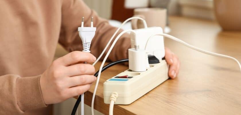 Uma pessoa está conectando um plugue branco de dois pinos em uma régua de energia bege que já possui vários outros cabos e carregadores conectados.