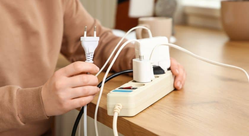 Uma pessoa está conectando um plugue branco de dois pinos em uma régua de energia bege que já possui vários outros cabos e carregadores conectados.