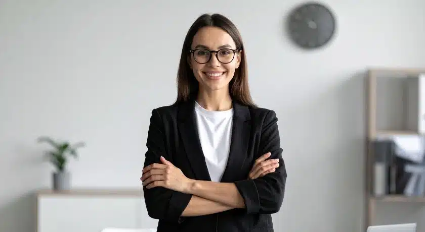 uma mulher de negócios sorridente, de pele clara e cabelos castanhos longos, em pé, olhando para frente. Ela usa óculos de aros pretos, uma camisa branca por baixo de um blazer preto, e está com os braços cruzados.
