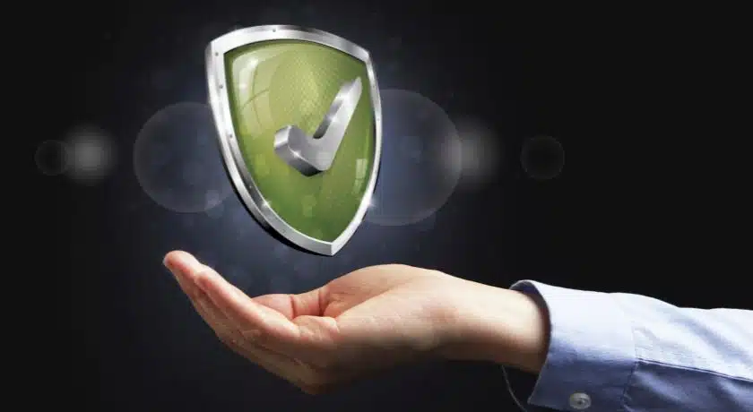 Uma mão aberta, vestindo uma camisa azul clara com as mangas arregaçadas, parece sustentar no ar um escudo tridimensional. O escudo é predominantemente verde-oliva com bordas e detalhes prateados metálicos, dando uma aparência robusta e tecnológica. No centro do escudo, há um grande "tick" (símbolo de verificação) prateado