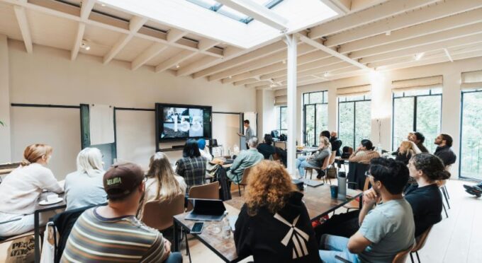 Uma reunião, treinamento ou apresentação acontecendo em uma sala ampla e bem iluminada. Cerca de 15 a 20 pessoas estão sentadas em cadeiras, e viradas para um instrutor que está em pé à frente. O instrutor está ao lado de uma grande TV de tela plana, que exibe um slide.
