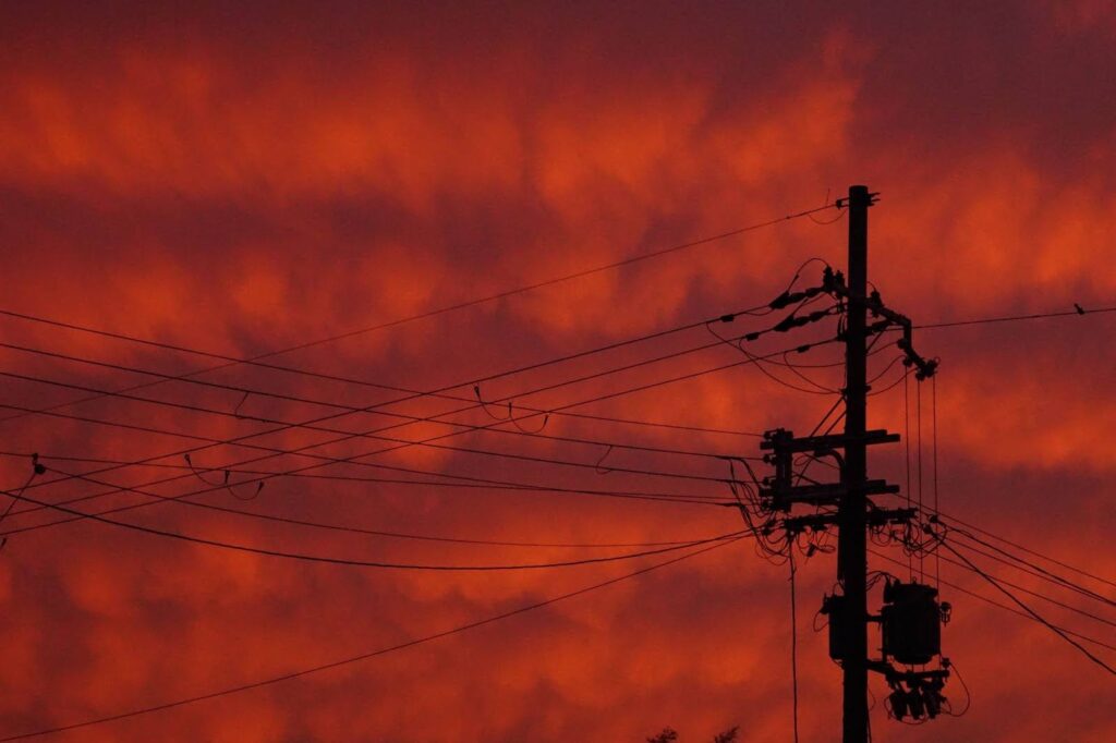 O céu está completamente tomado por um tom intenso de laranja-avermelhado, lembrando um pôr do sol dramático. No primeiro plano, há um poste de energia com vários fios cruzando a imagem em diferentes direções. Os cabos se entrelaçam formando um padrão geométrico contra o fundo vibrante do céu. A cena transmite uma sensação de calor e energia, com contraste forte entre as silhuetas escuras e o brilho do horizonte.