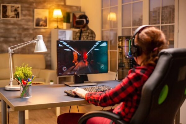 A imagem mostra uma mulher de costas, sentada em uma cadeira gamer preta e verde, usando headset e jogando em um desktop em uma sala de estar com iluminação aconchegante. Ela usa uma camisa xadrez vermelha e preta. À sua frente, um monitor exibe uma cena de jogo em primeira pessoa com gráficos de túnel e luzes vermelhas. Um gabinete de computador com luzes RGB está à direita do monitor, e uma luminária de mesa branca está à esquerda. No fundo desfocado, a sala tem paredes decoradas com fotos e abajures amarelos acesos.
