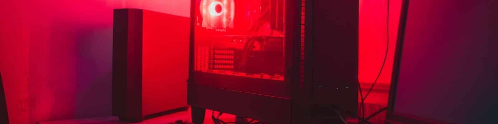Um computador de mesa com gabinete transparente iluminado por uma luz vermelha intensa. Dentro dele, é possível ver ventoinhas acesas e componentes internos. Ao lado, há um teclado, um mouse e um monitor apagado sobre uma mesa de superfície branca. Todo o ambiente está banhado por uma iluminação vermelha, criando um clima tecnológico e moderno.