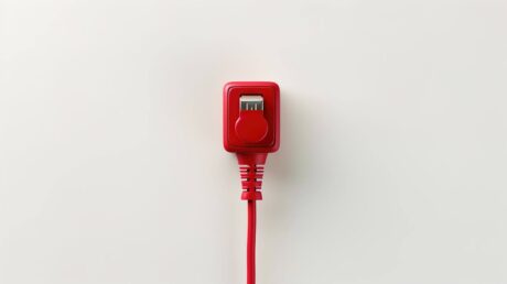 Esta é uma imagem de close-up de uma lâmpada incandescente ou LED com bulbo de vidro vermelho, deitada de lado sobre uma superfície escura e levemente texturizada. A lâmpada está acesa, emitindo uma luz vermelha intensa. Dentro do bulbo, os filamentos ou LEDs brilham em um tom amarelado/laranja forte. Um fio elétrico fino e vermelho sai da base metálica rosqueada da lâmpada, curvando-se para o lado. O brilho da lâmpada cria um forte halo vermelho na superfície circundante, sugerindo energia ou alerta.