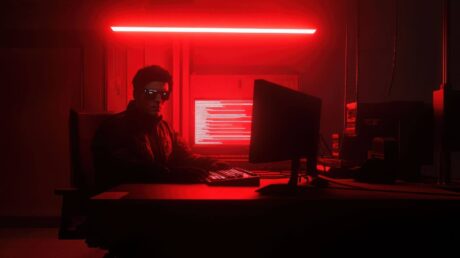 Uma mesa de escritório moderna está iluminada por uma luz vermelha suave. No centro, há um monitor grande e desligado, com um teclado e um mouse posicionados à frente. À esquerda, há uma pequena caixa de som preta, e à direita, uma luminária preta direciona a luz vermelha para a mesa. Também há uma caneca vermelha ao lado da luminária. O ambiente é escuro e minimalista, com destaque para o contraste entre o preto e o vermelho, criando uma atmosfera elegante e tecnológica.