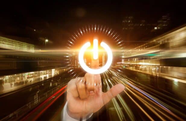 A imagem apresenta uma composição dinâmica que combina elementos reais e digitais. Em primeiro plano, uma mão (vestindo a manga de uma camisa branca) está estendida, com o dedo indicador tocando ou pairando sobre um grande e luminoso símbolo digital de "Power" (liga/desliga)