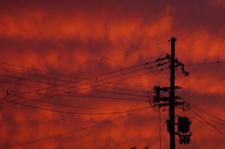 O céu está completamente tomado por um tom intenso de laranja-avermelhado, lembrando um pôr do sol dramático. No primeiro plano, há um poste de energia com vários fios cruzando a imagem em diferentes direções. Os cabos se entrelaçam formando um padrão geométrico contra o fundo vibrante do céu. A cena transmite uma sensação de calor e energia, com contraste forte entre as silhuetas escuras e o brilho do horizonte.