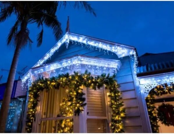 Fachada de uma casa decorada para o Natal ao anoitecer. O telhado está contornado com luzes brancas brilhantes no estilo cascata, e uma grande guirlanda verde com luzes amarelas envolve a janela principal.