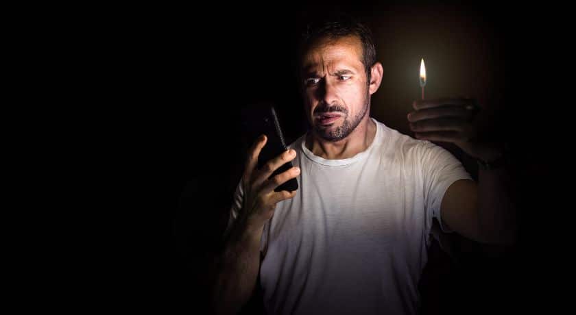 Fotografia de um homem em um ambiente completamente escuro, iluminado apenas pela luz de um palito de fósforo que ele segura em uma das mãos e pelo brilho da tela de um celular na outra. Ele tem uma expressão de preocupação e incerteza.