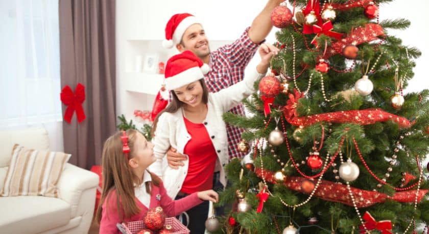 Uma família de três pessoas, incluindo um pai, uma mãe e uma filha, sorrindo enquanto decoram uma grande árvore de Natal na sala de estar. Todos usam gorros vermelhos de Papai Noel.