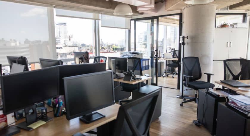 um escritório moderno e luminoso, com várias estações de trabalho vazias equipadas com computadores e cadeiras de escritório.