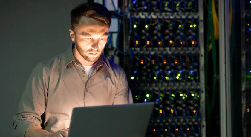 Homem, de barba e camisa clara, sentado em frente a um laptop em um ambiente escuro, iluminado pela luz da tela do computador e pelo brilho de várias luzes de LED verdes e amarelas vindas de racks de servidores atrás dele.
