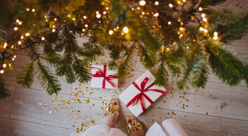 Visão em primeira pessoa de presentes embrulhados em papel branco com laços vermelhos, colocados sob uma árvore de Natal iluminada com luzes amarelas. Confetes dourados estão espalhados no chão de madeira clara, e os pés de uma pessoa usando sapatos dourados brilhantes estão visíveis ao lado dos presentes