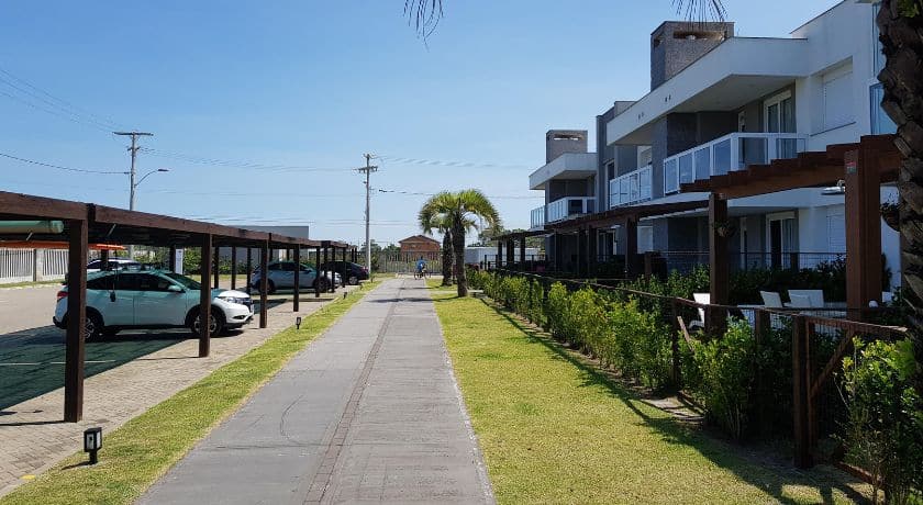Vista de uma passarela de concreto em um condomínio ou complexo de apartamentos. Há vagas de estacionamento cobertas por pergolados de madeira à esquerda e edifícios de dois andares com sacadas e paisagismo à direita.