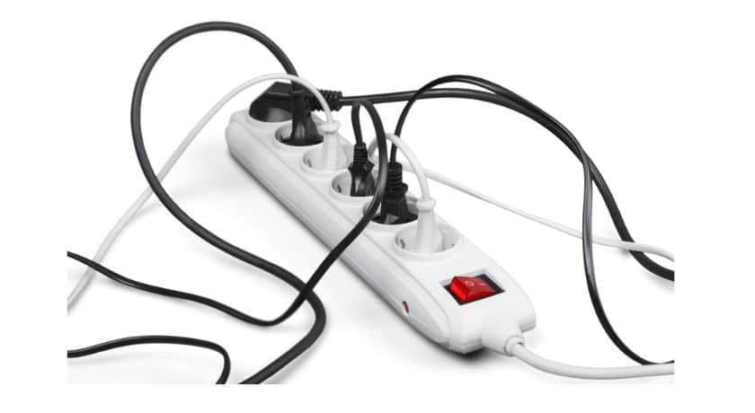 Uma régua de energia branca comum (extensão múltipla) com três tomadas e um interruptor liga/desliga vermelho está em foco. Vários cabos pretos e um cabo branco estão conectados às tomadas da régua, criando uma confusão de fios emaranhados.