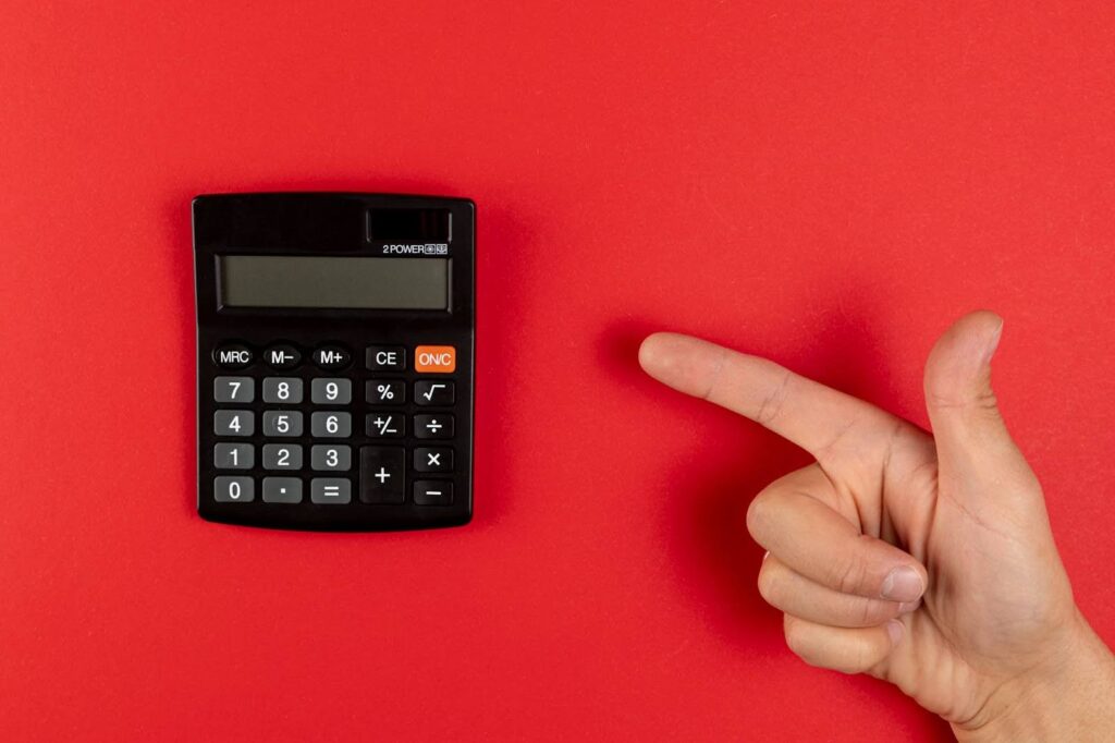 Uma foto de ângulo superior (flat lay) mostra uma calculadora de bolso preta à esquerda e uma mão com o dedo indicador esticado à direita, ambas em um fundo vermelho sólido e vibrante.