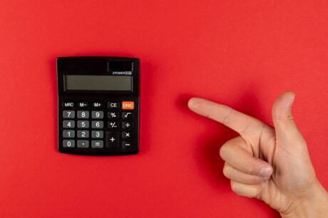 Uma foto de ângulo superior (flat lay) mostra uma calculadora de bolso preta à esquerda e uma mão com o dedo indicador esticado à direita, ambas em um fundo vermelho sólido e vibrante.