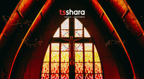 Esta imagem mostra o interior de um ambiente religioso, provavelmente uma igreja, dominado por uma iluminação dramática em tons de vermelho e laranja. O foco está no grande vitral colorido, que preenche uma parede. O vitral tem um padrão geométrico, em tons fortes de amarelo e vermelho fogo, e é emoldurado por uma estrutura angular no teto. No centro do vitral, um crucifixo com a figura de Cristo está suspenso. No topo da imagem, sobre a estrutura escura do teto abobadado, está o logotipo "ts shara" em vermelho, com o texto "nobreaks | estabilizadores" abaixo.