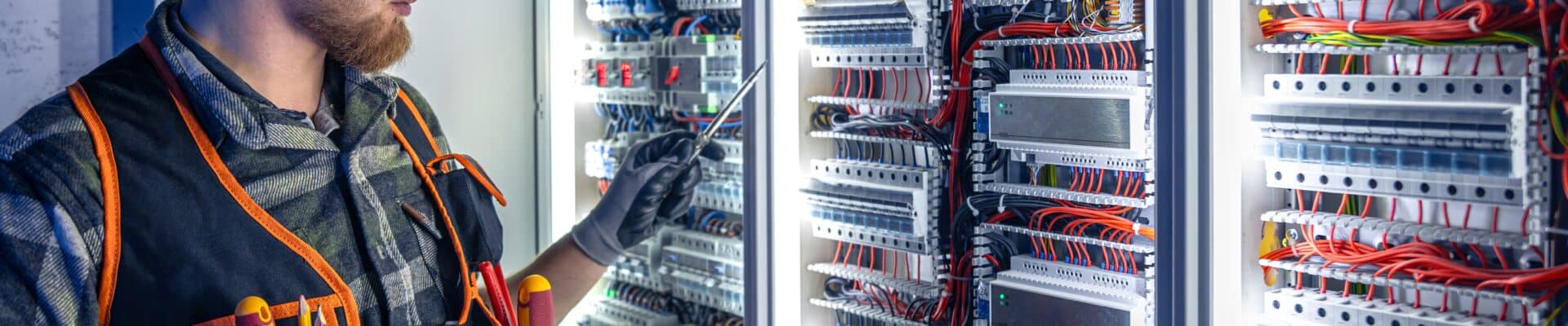 Um eletricista especializado, usando capacete branco com lanterna de cabeça, óculos de proteção e colete de ferramentas, trabalha em frente a um grande painel elétrico industrial aberto.