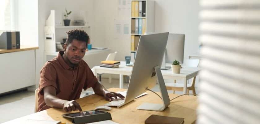 um homem negro jovem trabalhando em um escritório moderno, bem iluminado e organizado. Ele está sentado à mesa de madeira, concentrado, enquanto utiliza um tablet com a mão esquerda e tem um computador desktop à sua frente