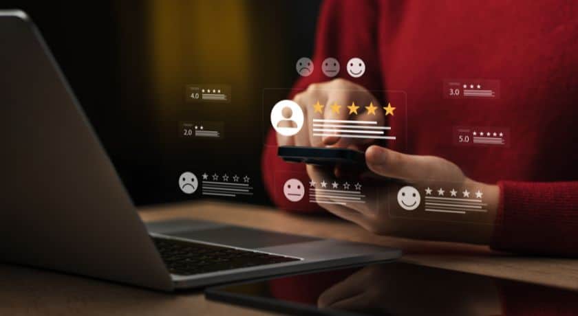 Uma pessoa (usando uma camisa vermelha, visível apenas do torso e braços) está sentada em uma mesa escura com um laptop aberto. Ela segura um smartphone e interage com uma interface digital que projeta ícones de avaliação e feedback no ar. A interface mostra perfis de usuário, estrelas de avaliação (por exemplo, 2.0, 3.0, 4.0, e um perfil central com 4 de 5 estrelas