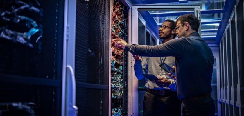 Dois técnicos de TI (Tecnologia da Informação), um com óculos, estão lado a lado em um corredor iluminado por luzes azuis de um centro de dados (Data Center). Eles estão inspecionando e apontando para os cabos e equipamentos dentro de um rack de servidores aberto.