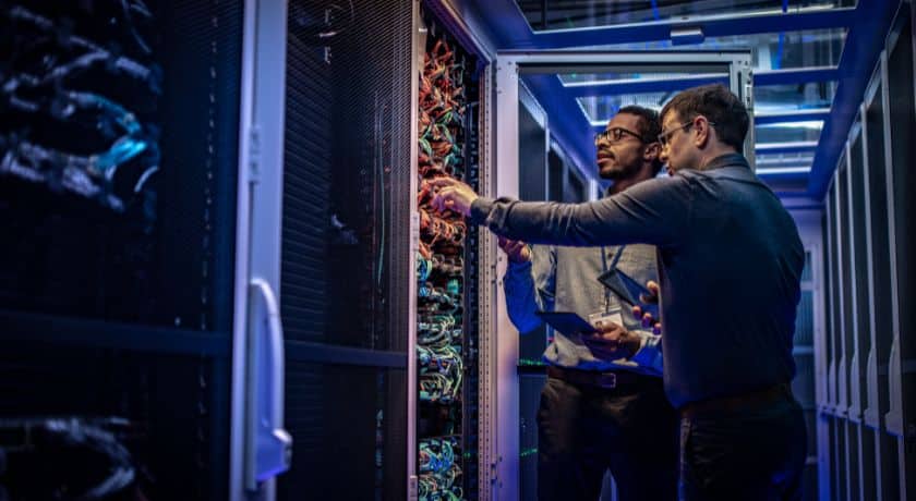 Dois técnicos de TI (Tecnologia da Informação), um com óculos, estão lado a lado em um corredor iluminado por luzes azuis de um centro de dados (Data Center). Eles estão inspecionando e apontando para os cabos e equipamentos dentro de um rack de servidores aberto.
