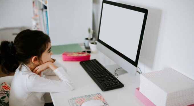 Uma criança vista de perfil, sentada em uma escrivaninha branca, olhando atentamente para a tela de um computador desktop. O ambiente sugere um momento de estudo ou aula remota em casa