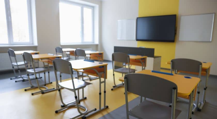 Interior de uma sala de aula moderna e iluminada, com várias mesas e cadeiras escolares amarelas e cinzas organizadas em fileiras. Ao fundo, uma parede com detalhe amarelo exibe uma TV de tela plana grande