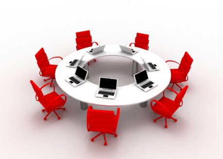 Mesa de reunião branca em formato circular, com abertura central, rodeada por cadeiras giratórias vermelhas. Sobre a mesa, vários notebooks abertos estão distribuídos de forma uniforme, sugerindo um ambiente corporativo ou colaborativo. Fundo claro e neutro.