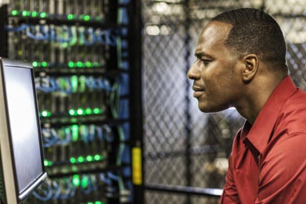 Retrato de perfil de um técnico negro, vestindo uma camisa social vermelha, concentrado enquanto olha para um monitor de computador em um centro de dados. Ao fundo, de forma desfocada, observa-se um rack de servidores repleto de luzes LED verdes brilhantes e diversos cabos de rede azuis organizados, simbolizando um ambiente de infraestrutura tecnológica complexo e ativo.