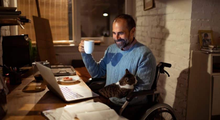 Homem em cadeira de rodas trabalhando em home office à noite, sorrindo com uma caneca na mão e um gato no colo enquanto olha para o laptop.