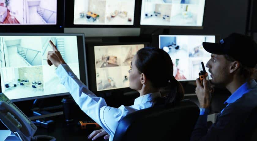 um homem e uma mulher uniformizados em uma sala apontando para cameras de monitoramento de um estabelecimento comercial
