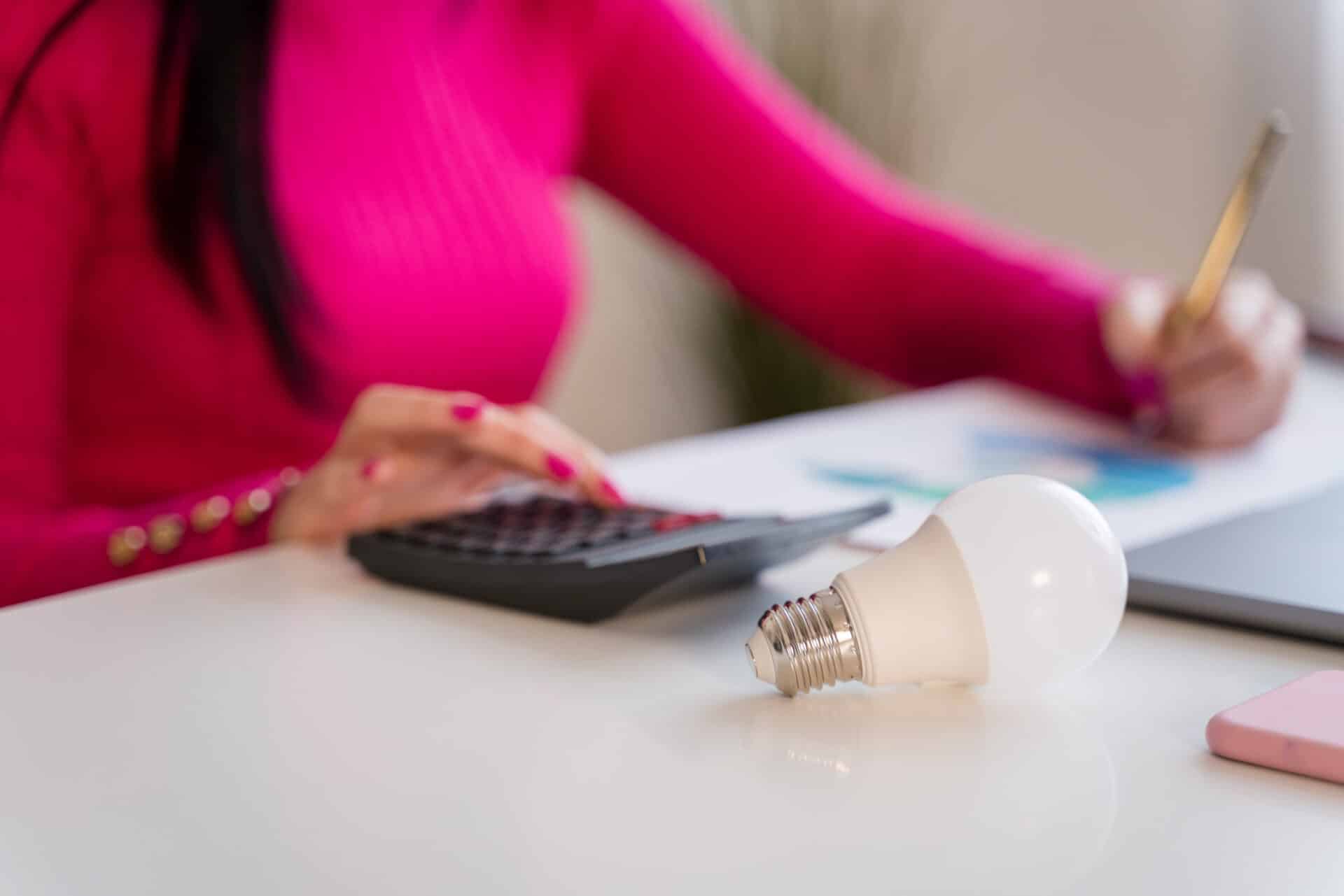 Lâmpada sobre mesa enquanto pessoa usa calculadora e anota gastos de energia.