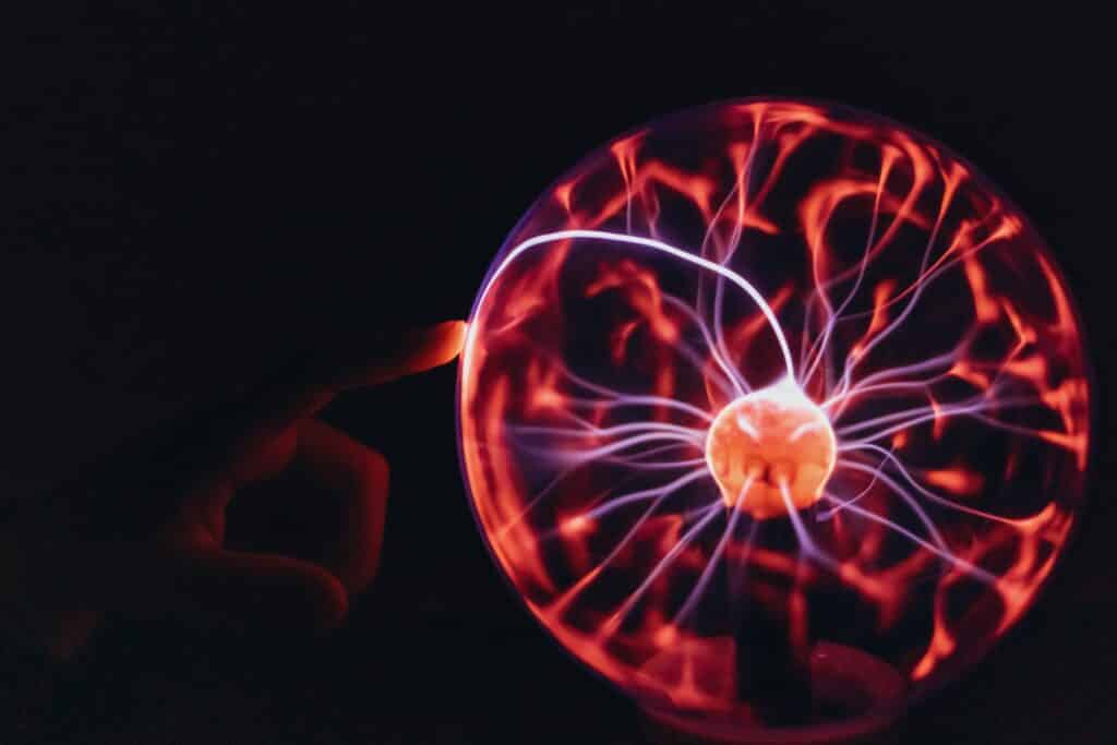 um globo de plasma em um ambiente escuro. No centro da esfera de vidro, um núcleo brilhante emite filamentos de eletricidade em tons de rosa e roxo que se estendem até a borda. Um dedo toca a superfície externa do vidro, fazendo com que um feixe de luz mais intenso e concentrado seja atraído para o ponto de contato, ilustrando visualmente a condução de energia estática.