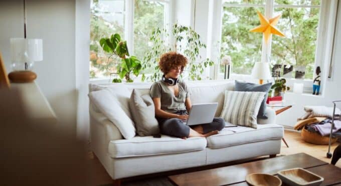 Uma mulher com cabelos cacheados e volumosos, usando fones de ouvido ao redor do pescoço, senta-se no sofá com pernas cruzadas, focada em seu laptop.