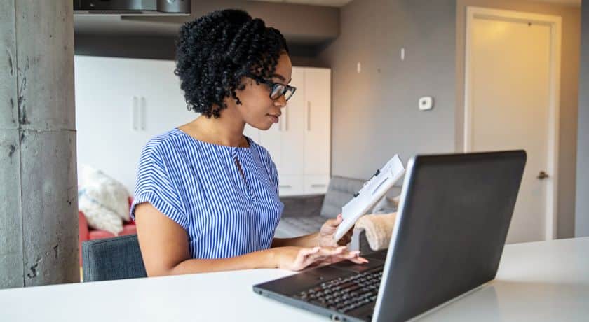 uma mulher sentada em uma cadeira na sala de estar com o notebook aberto e segurando um livro