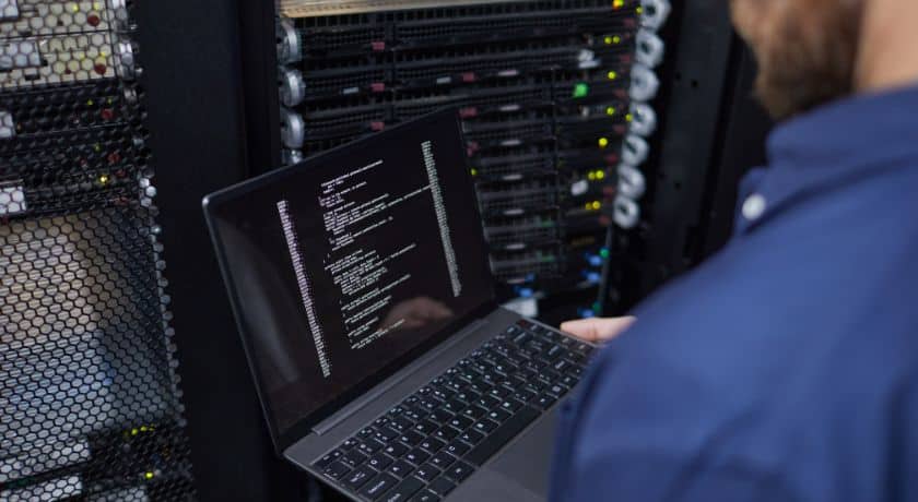 Vista traseira de um profissional de TI segurando um notebook aberto que exibe linhas de código, com racks de servidores de data center