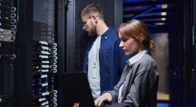 Dois profissionais de TI trabalhando em uma sala de servidores climatizada; a mulher em primeiro plano utiliza um laptop para monitorar o sistema