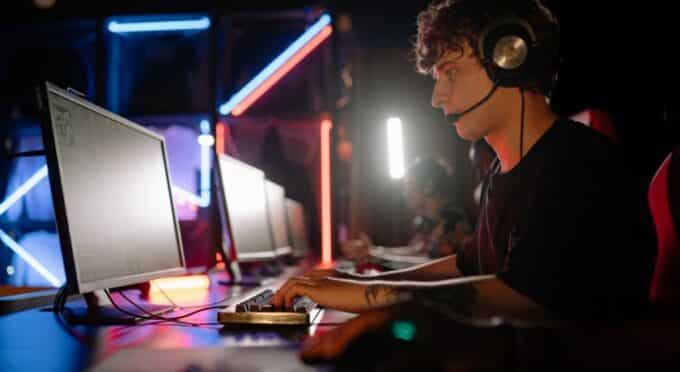 Um jovem de perfil, usando fones de ouvido com microfone, joga em um computador em uma arena de e-sports.