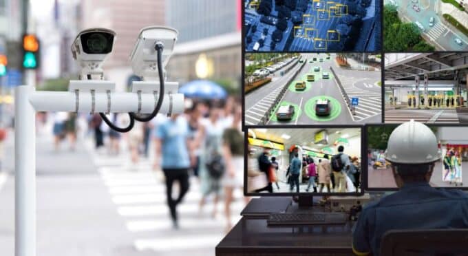 Uma imagem composta dividida em duas partes que ilustra a vigilância moderna. À esquerda, em destaque, duas câmeras de segurança brancas estão montadas em um poste, voltadas para uma rua movimentada da cidade com pedestres desfocados ao fundo. À direita, uma visão de um centro de monitoramento mostra um operador de capacete branco de costas, observando vários monitores