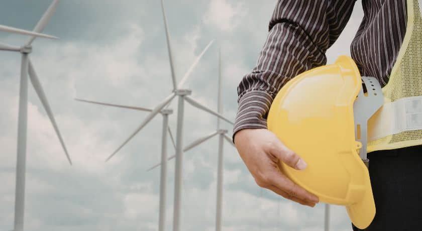 Close lateral de um profissional de engenharia segurando um capacete de segurança amarelo sob o braço, com um parque eólico ao fundo sob um céu nublado.