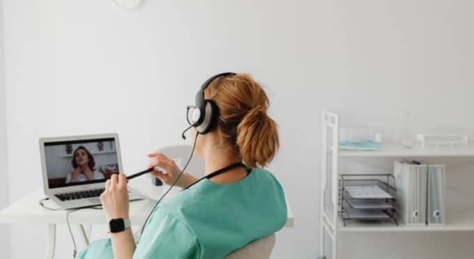 Vista traseira de uma profissional de saúde usando uniforme verde e headset, realizando uma consulta de telemedicina por meio de um notebook em um consultório médico